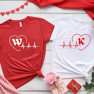 Pode incluir: Duas camisetas, uma vermelha e outra branca, com desenhos em forma de coração. A camiseta vermelha tem um "W" branco dentro de um coração com uma linha de batimento cardíaco. A camiseta branca tem um "K" vermelho dentro de um coração. Tema do Dia dos Namorados.