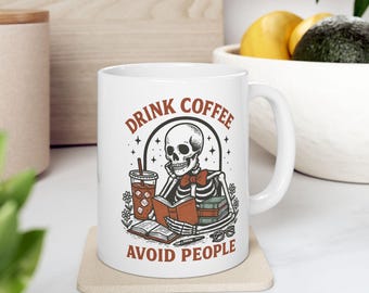 Könnte beinhalten: Weiße Keramik-Tasse mit schwarzer und roter Grafik. Die Grafik zeigt ein Skelett, das ein Buch liest, eine Tasse Kaffee und den Text "DRINK COFFEE AVOID PEOPLE". Die Tasse hat einen gebogenen Henkel und steht auf einem beigen Untersetzer.