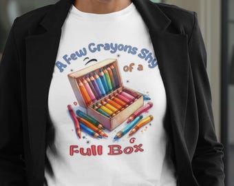 Könnte beinhalten: Weißes T-Shirt mit einer farbenfrohen Illustration einer Buntstiftbox und dem Text "A Few Crayons Shy of a Full Box". Das Design umfasst Buntstifte in verschiedenen Farben.