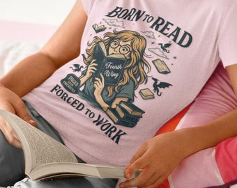 May include: A light pink t-shirt featuring a graphic illustration of a person with glasses reading a book titled Fourth Wing. The text reads BORN TO READ and FORCED TO WORK, with additional small drawings of dragons, clouds, and books surrounding the central figure.