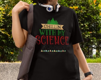 Pode incluir: Uma t-shirt preta de manga curta com uma impressão gráfica de pinheiros verdes e o texto "CHILLIN WITH MY SCIENCE SNOWMIES" em letras douradas, verdes e vermelhas, acompanhado por uma fila de pequenos ícones coloridos na parte inferior.