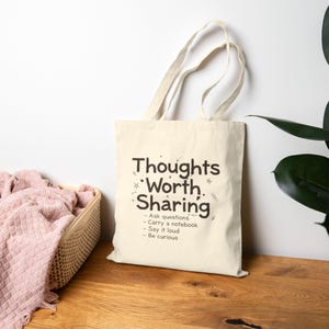 Puede incluir: Un bolso tote de color crema con las palabras "Thoughts Worth Sharing" en negro. La bolsa tiene una lista de indicaciones debajo del título. La bolsa tiene dos asas y está sobre una superficie de madera.
