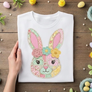 Op de afbeelding: Wit T-shirt met een kleurrijk konijnengezicht ontwerp. Het konijn heeft roze oren, hartvormige brillen en een bloemenkroon. Het ontwerp is gemaakt van patchwork stof en is omgeven door decoratieve paaseieren en kuikens.