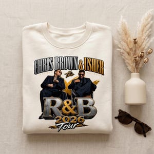 May include: A cream-coloured sweatshirt with a graphic of Chris Brown and Usher. The design includes the text "CHRIS BROWN & USHER" above "The R&B 2026 Tour" in a stylized font. The image shows the two artists in suits.