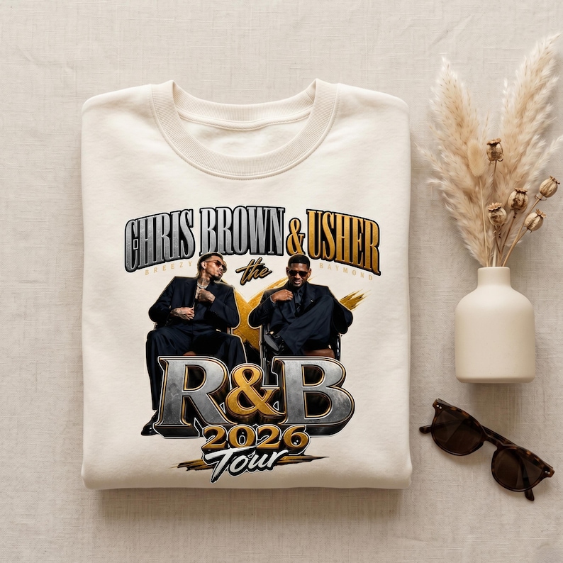 May include: A cream-coloured sweatshirt with a graphic of Chris Brown and Usher. The design includes the text "CHRIS BROWN & USHER" above "The R&B 2026 Tour" in a stylized font. The image shows the two artists in suits.