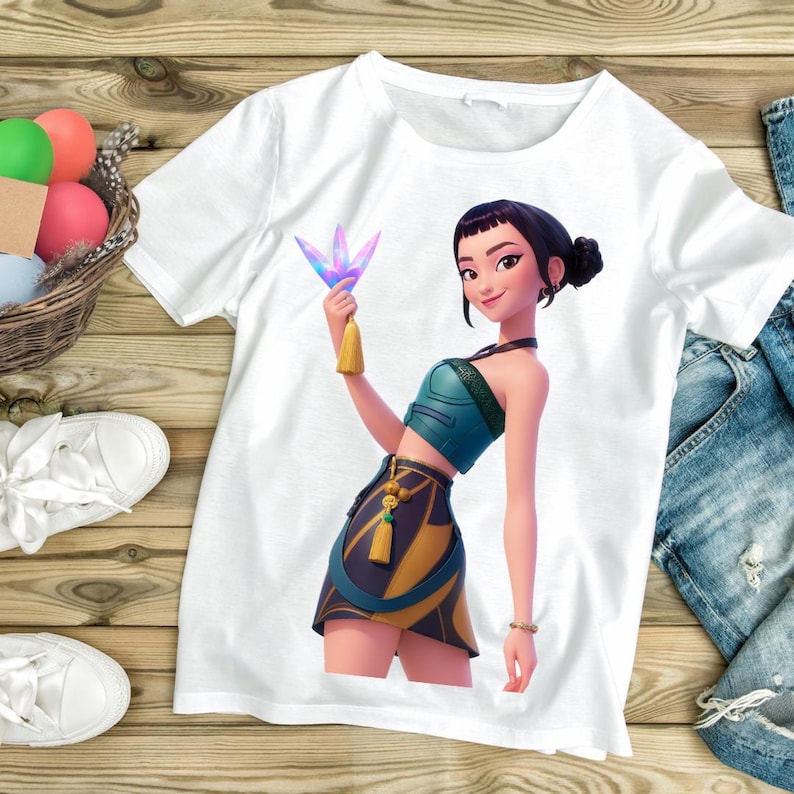May include: White t-shirt featuring a cartoon illustration of a woman in a teal top and skirt, holding a colorful crystal. The woman has black hair and is wearing a bracelet. The shirt is on a wooden surface.