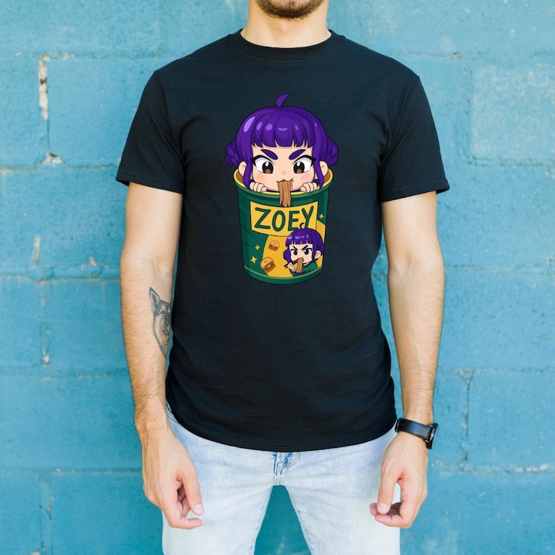 May include: Black t-shirt featuring a cartoon graphic of a purple-haired character in a green cup with the name "ZOEY". The design includes a smaller version of the character and the cup. The shirt is worn by a person.