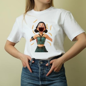 May include: White t-shirt featuring a cartoon illustration of a woman with an open mouth and orange accents. The woman is wearing a teal top and dark green pants. The shirt is made of a soft, comfortable material.