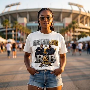 May include: White t-shirt with the text "CHRIS BROWN & USHER" above a graphic of two men in suits. Below the graphic is "R&B 2026 TOUR" in gold and silver lettering. The shirt is worn by a person in blue denim shorts.