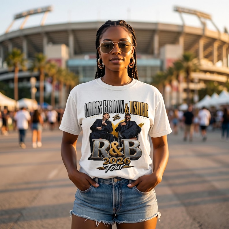 May include: White t-shirt with the text "CHRIS BROWN & USHER" above a graphic of two men in suits. Below the graphic is "R&B 2026 TOUR" in gold and silver lettering. The shirt is worn by a person in blue denim shorts.