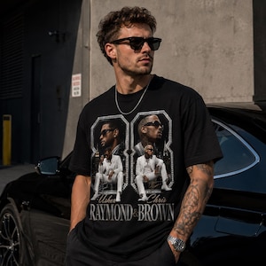 K&ouml;nnte beinhalten: Ein schwarzes Kurzarm-T-Shirt mit einem Grafikdruck von Usher und Chris Brown in wei&szlig;en Anz&uuml;gen. Das Design enth&auml;lt zwei gerahmte Portr&auml;ts und den Text "RAYMOND & BROWN" in einer fetten, metallisch wirkenden Schrift auf der Brust.