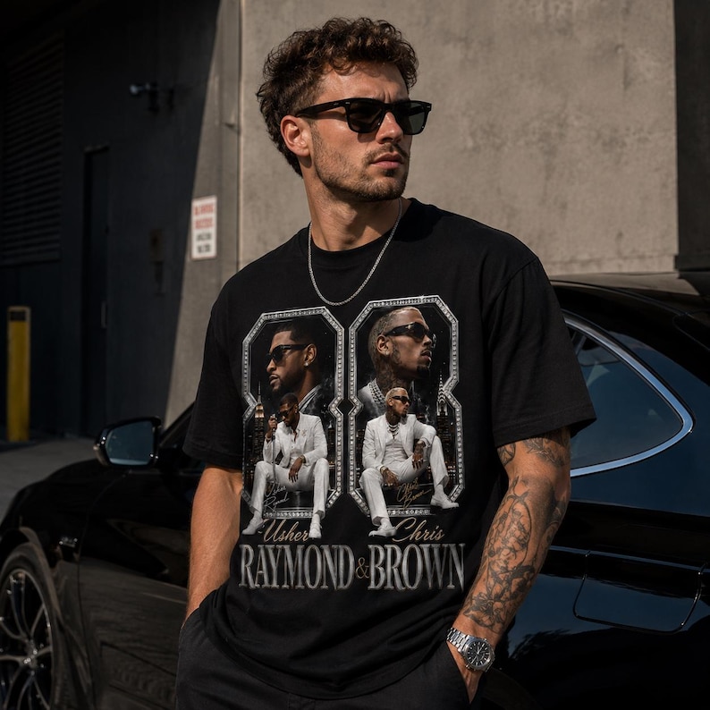 K&ouml;nnte beinhalten: Ein schwarzes Kurzarm-T-Shirt mit einem Grafikdruck von Usher und Chris Brown in wei&szlig;en Anz&uuml;gen. Das Design enth&auml;lt zwei gerahmte Portr&auml;ts und den Text "RAYMOND & BROWN" in einer fetten, metallisch wirkenden Schrift auf der Brust.