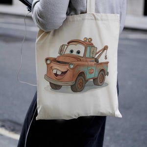 May include: A beige canvas tote bag featuring a cartoon illustration of Mater from the movie Cars. The image shows Mater in shades of brown, blue, and white, with a smiling face and a tow hook. The bag is held by a person.