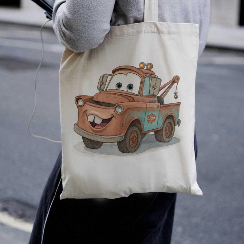 May include: A beige canvas tote bag featuring a cartoon illustration of Mater from the movie Cars. The image shows Mater in shades of brown, blue, and white, with a smiling face and a tow hook. The bag is held by a person.