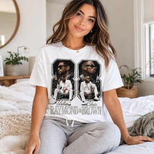K&ouml;nnte beinhalten: Ein wei&szlig;es Kurzarm-T-Shirt mit einem Grafikdruck von zwei M&auml;nnern in wei&szlig;en Anz&uuml;gen vor einer Skyline. Das Design enth&auml;lt den Text Usher RAYMOND & BROWN in einer stilisierten Schriftart, eingerahmt von zwei gro&szlig;en Ziffern in Form einer Acht.