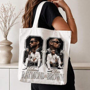 K&ouml;nnte beinhalten: Eine wei&szlig;e Stofftasche mit einem Grafikdruck von zwei M&auml;nnern in wei&szlig;en Anz&uuml;gen und Sonnenbrillen, die jeweils in einer gro&szlig;en, mit Diamanten besetzten Acht eingerahmt sind. Der Text "Usher RAYMOND & BROWN" ist unten in silberner Schrift gedruckt.