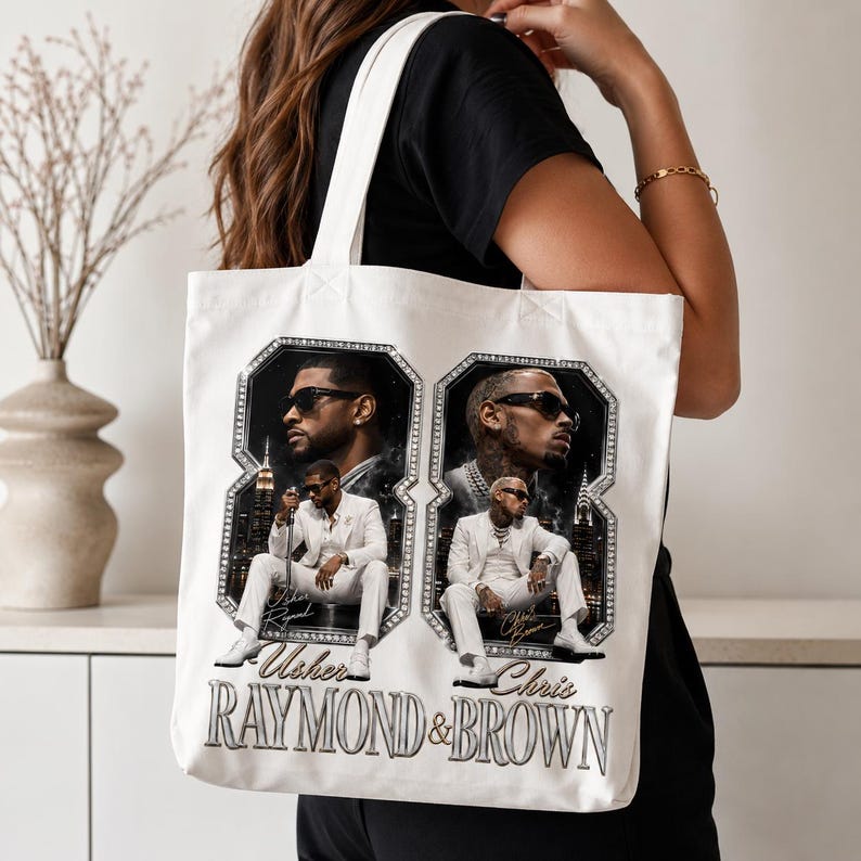 K&ouml;nnte beinhalten: Eine wei&szlig;e Stofftasche mit einem Grafikdruck von zwei M&auml;nnern in wei&szlig;en Anz&uuml;gen und Sonnenbrillen, die jeweils in einer gro&szlig;en, mit Diamanten besetzten Acht eingerahmt sind. Der Text "Usher RAYMOND & BROWN" ist unten in silberner Schrift gedruckt.