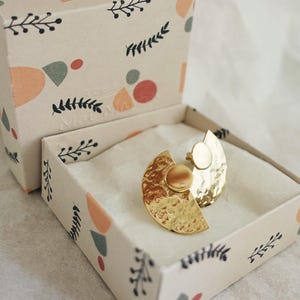 May include: A pair of gold-toned earrings with a hammered texture and a semi-circular shape. The earrings are in a small, white box with a floral pattern.