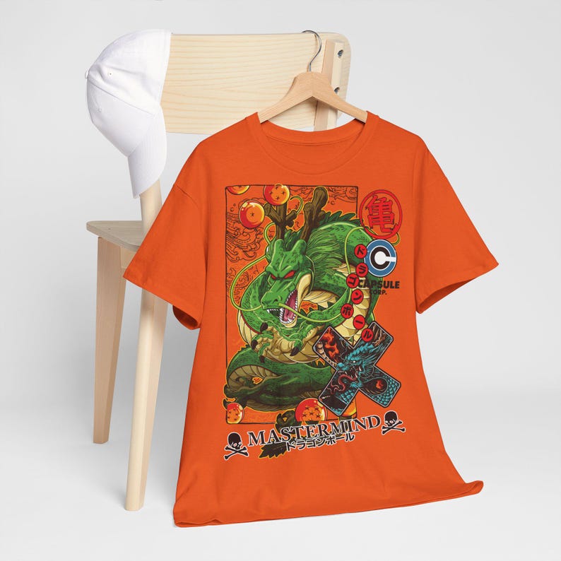 May include: Orange t-shirt featuring a green dragon illustration with the text "MASTERMIND" and Japanese text. The design includes dragon balls and other graphic elements. A white baseball cap rests on a wooden chair.