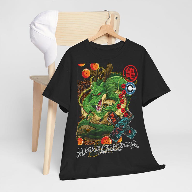 May include: Black t-shirt featuring a green dragon illustration with orange orbs and Japanese text. The design includes the text "MASTERMIND" and "ドラゴンボール". A white baseball cap hangs on a wooden chair in the background.