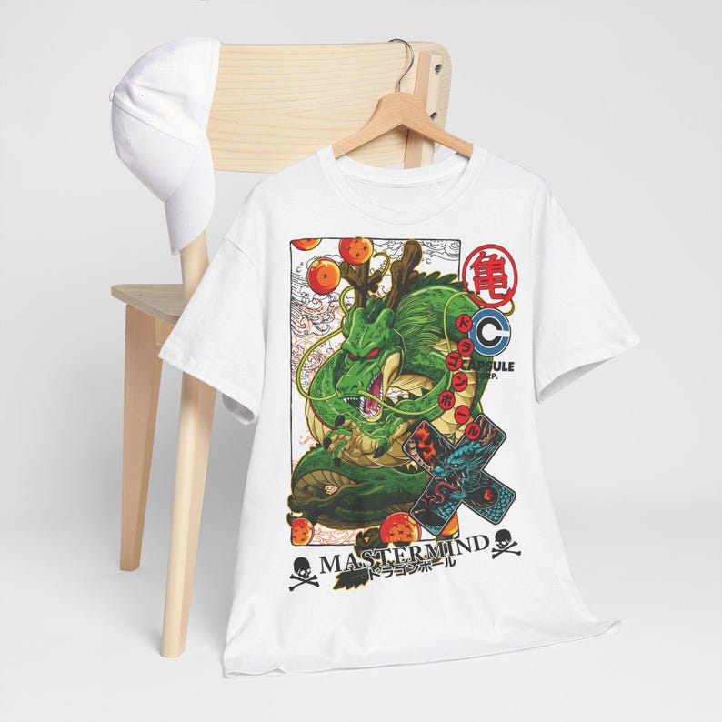 May include: White t-shirt featuring a green dragon illustration with orange orbs and Japanese text. The design includes the words "MASTERMIND" and "CAPSULE". A white baseball cap hangs on a wooden chair.