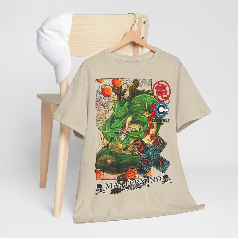 May include: Beige t-shirt featuring a green dragon illustration with orange orbs and Japanese text. The design includes the text "MASTERMIND" and "CAPSULE CORP." along with skull and crossbones symbols. A white baseball cap rests on a wooden chair.