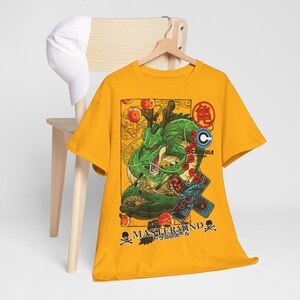 May include: Mustard yellow t-shirt featuring a green dragon illustration with orange orbs and Japanese text. The shirt also has the text "MASTERMIND" and a skull and crossbones graphic. A white baseball cap rests on a wooden chair.