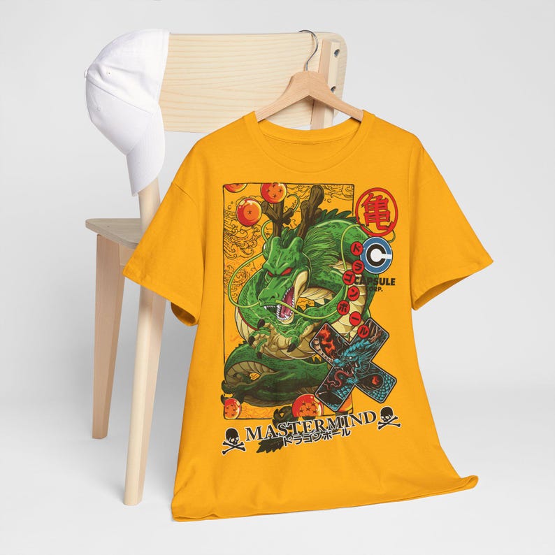 May include: Mustard yellow t-shirt featuring a green dragon illustration with orange orbs and Japanese text. The shirt also has the text "MASTERMIND" and a skull and crossbones graphic. A white baseball cap rests on a wooden chair.