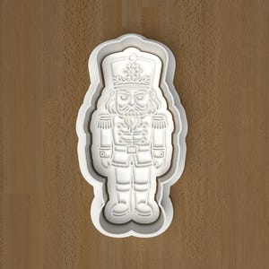 May include: A white plastic cookie cutter in the shape of a nutcracker soldier. The cutter has detailed features including a hat, uniform, and facial details. The cutter is on a brown wooden surface.