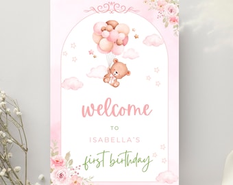 May include: A pink and white welcome sign for a first birthday. The sign features a teddy bear floating with balloons, clouds, and stars. The text reads "welcome to Isabella's first birthday" in pink and green.