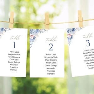 May include: Three white table cards with floral accents and the word "Table" at the top. Each card has a number and guest names. The cards are hanging from a string with wooden clothespins.