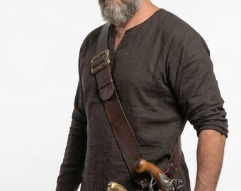 May include: A brown leather shoulder belt with a brass buckle holds a flintlock pistol and a sabre. The pistol has a wooden handle. The sabre has a gold-coloured hilt. The person is wearing a brown long-sleeved shirt.
