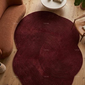 Può includere: Un tappeto bordeaux dalla forma astratta con una superficie strutturata e scanalata. Il design del tappeto presenta motivi a spirale sovrapposti. È posto su un pavimento in legno, con un divano e un tavolo sullo sfondo.