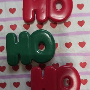 May include: Three plastic "HO" Christmas ornaments in red and green. The letters are on a white background with red heart shapes and pink stripes.