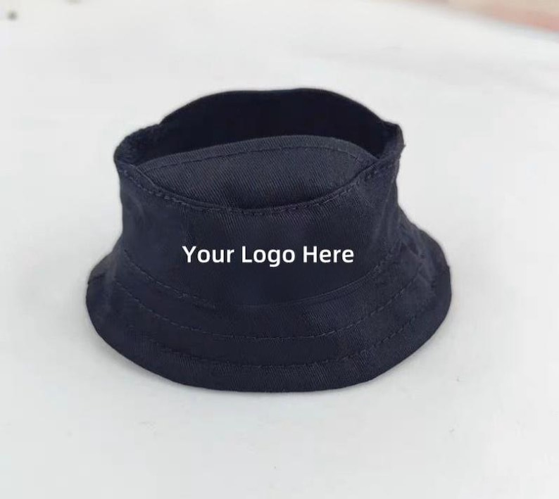 K&ouml;nnte beinhalten: Ein marineblauer Fischerhut mit dem Text "Your Logo Here" in Wei&szlig;. Der Hut hat eine runde Krone und eine nach unten geneigte Krempe. Der Hut ist aus einem robusten Stoff gefertigt.