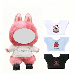 Può includere: Una bambola di coniglio rosa di peluche con una maglietta bianca con la scritta "Your Logo Here". Tre magliette in miniatura con grafiche: una maglietta bianca con un design "Cancelled Forever", una maglietta blu con una mela e una maglietta nera con un orso.