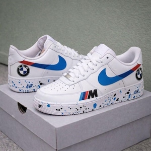 Könnte beinhalten: Weiße Turnschuhe mit blauen Nike-Swooshes und Akzenten. Die Schuhe haben BMW- und M-Logos mit blauen, roten und schwarzen Details. Die Sohlen haben ein gesprenkeltes Farbmuster mit dem Wort "AIR".