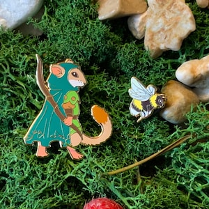 Puede incluir: Un pin de esmalte dorado con un roedor de dibujos animados que lleva una capa con capucha verde y azul y sostiene un arco y una flecha. El roedor tiene una cola larga y está de pie sobre una superficie verde musgosa. También se puede ver un pequeño pin de esmalte dorado de una abeja en la imagen.