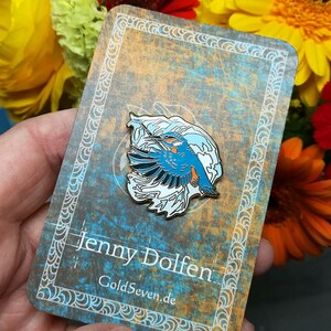 May include: A blue and white enamel pin featuring a kingfisher bird in flight with a wave design. The pin is on a card with the text "Jenny Dolfen" and "GoldSeven.de".