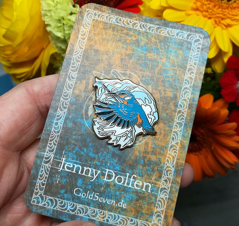 May include: A blue and white enamel pin featuring a kingfisher bird in flight with a wave design. The pin is on a card with the text "Jenny Dolfen" and "GoldSeven.de".