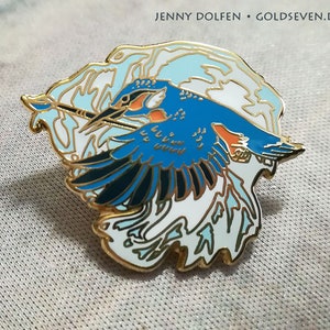 May include: A gold-toned enamel pin featuring a blue and orange kingfisher bird in flight with a paintbrush in its beak. The bird is surrounded by a white and blue abstract design.