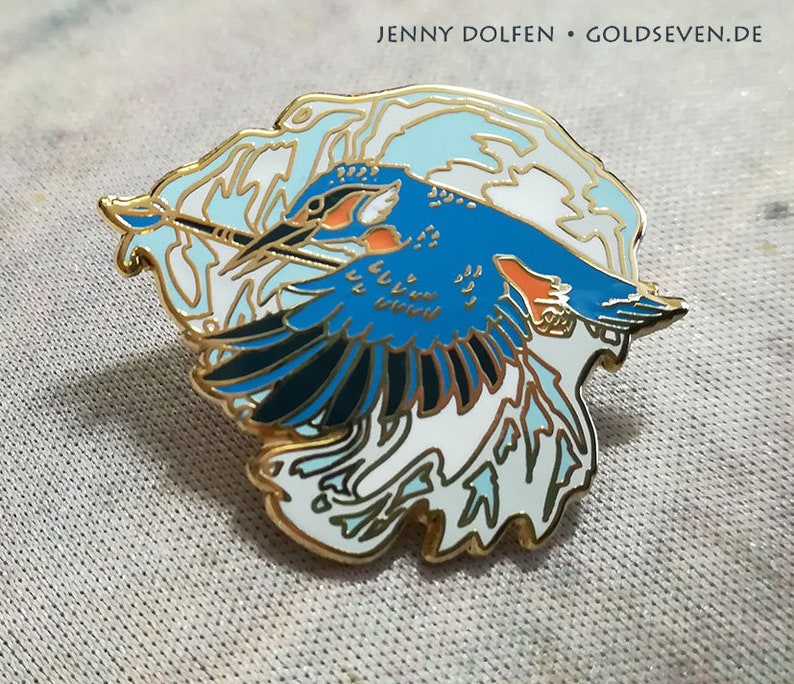 May include: A gold-toned enamel pin featuring a blue and orange kingfisher bird in flight with a paintbrush in its beak. The bird is surrounded by a white and blue abstract design.