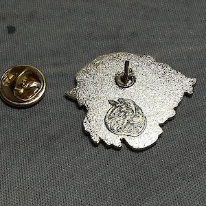 May include: The back of a silver metal pin with a detailed design. The pin has a safety clasp on the back.