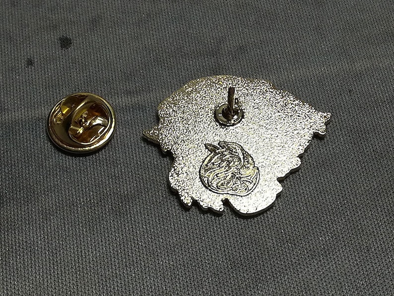 May include: The back of a silver metal pin with a detailed design. The pin has a safety clasp on the back.