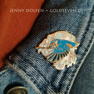 May include: Gold enamel pin featuring a blue kingfisher bird in flight with white and pink clouds. The pin is on a blue denim jacket with the text "JENNY DOLFEN &bull; GOLDSEVEN.DE".