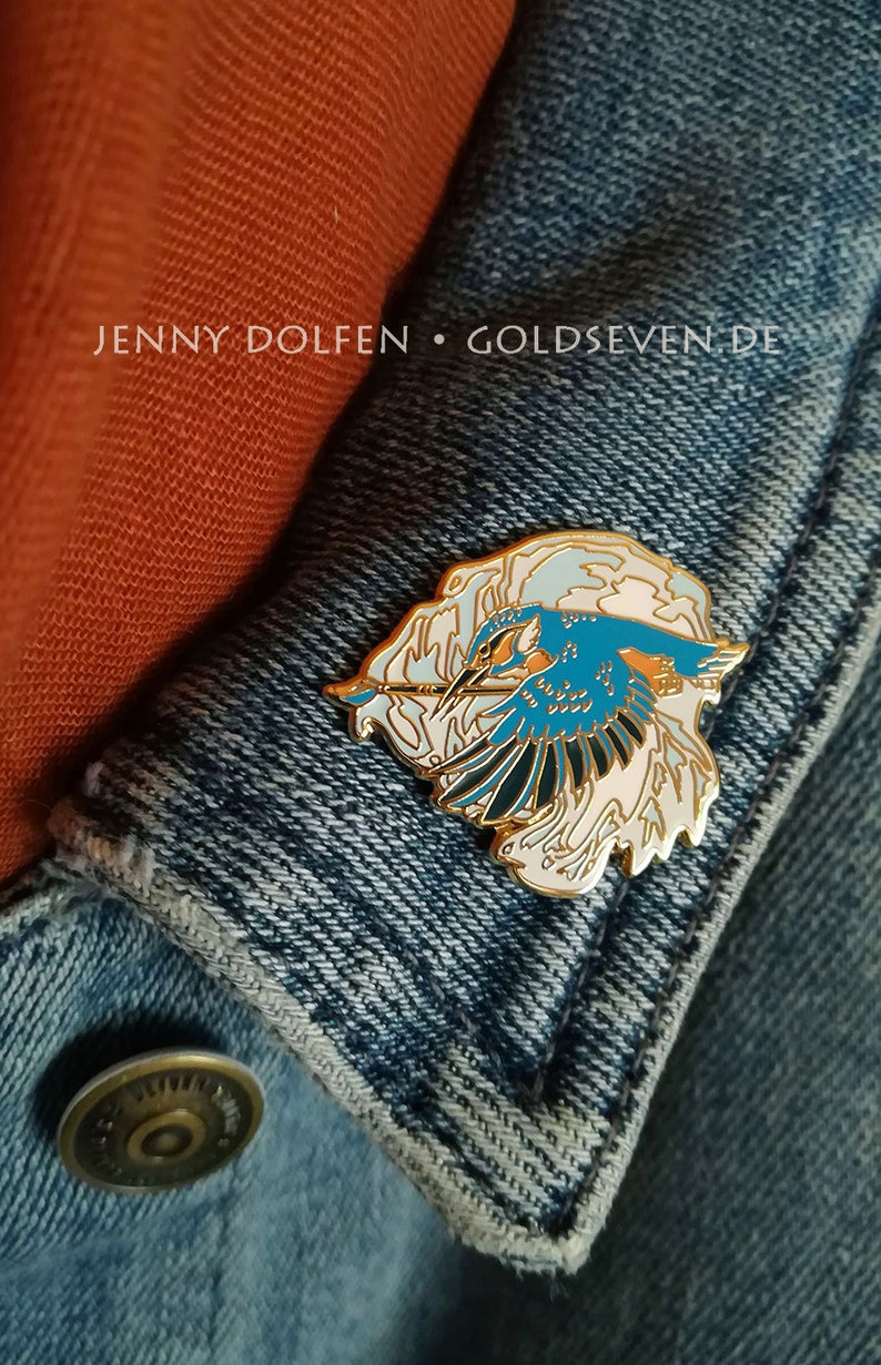 May include: Gold enamel pin featuring a blue kingfisher bird in flight with white and pink clouds. The pin is on a blue denim jacket with the text "JENNY DOLFEN &bull; GOLDSEVEN.DE".