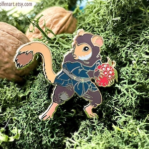 May include: A gold-toned enamel pin featuring a cartoon depiction of a small, furry creature wearing a blue and brown hooded cloak and holding a cluster of red berries. The creature has a long, bushy tail and a friendly expression.