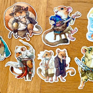 Op de afbeelding: Een verzameling grillige, geïllustreerde stickers met antropomorfe dieren. De stickers bevatten een konijn met een cape, een muis met een luit en een kat met een tovenaars hoed. Het kleurenpalet is voornamelijk op waterverf gebaseerd.