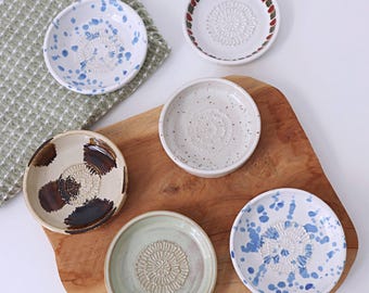 Może przedstawiać: Zestaw sześciu małych ceramicznych talerzy do ścierania w różnych kolorach i wzorach. Talerze ułożone są na drewnianej desce do krojenia i zielonej ściereczce o strukturze wafla. Każdy talerz posiada centralną powierzchnię do ścierania.