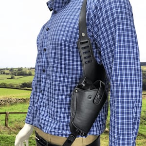 Puede incluir: Funda de hombro de cuero negro con una correa vertical y una funda para una pistola. La funda se lleva sobre una camisa a cuadros azul. El fondo muestra un campo verde y un cielo azul.